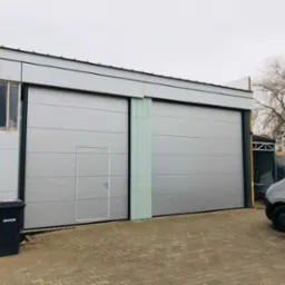 Szara brama przemysłowa z drzwiami w Tychach, widoczna z zewnątrz budynku. Obok zaparkowany srebrny bus i czarny kosz na śmieci.