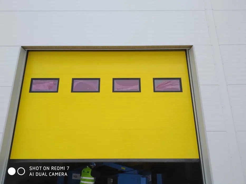 Żółta brama segmentowa z czterema okienkami, zamontowana w białej ścianie hali, widoczny fragment maszyny i osoba w odblaskowej kamizelce w dolnej części kadru.