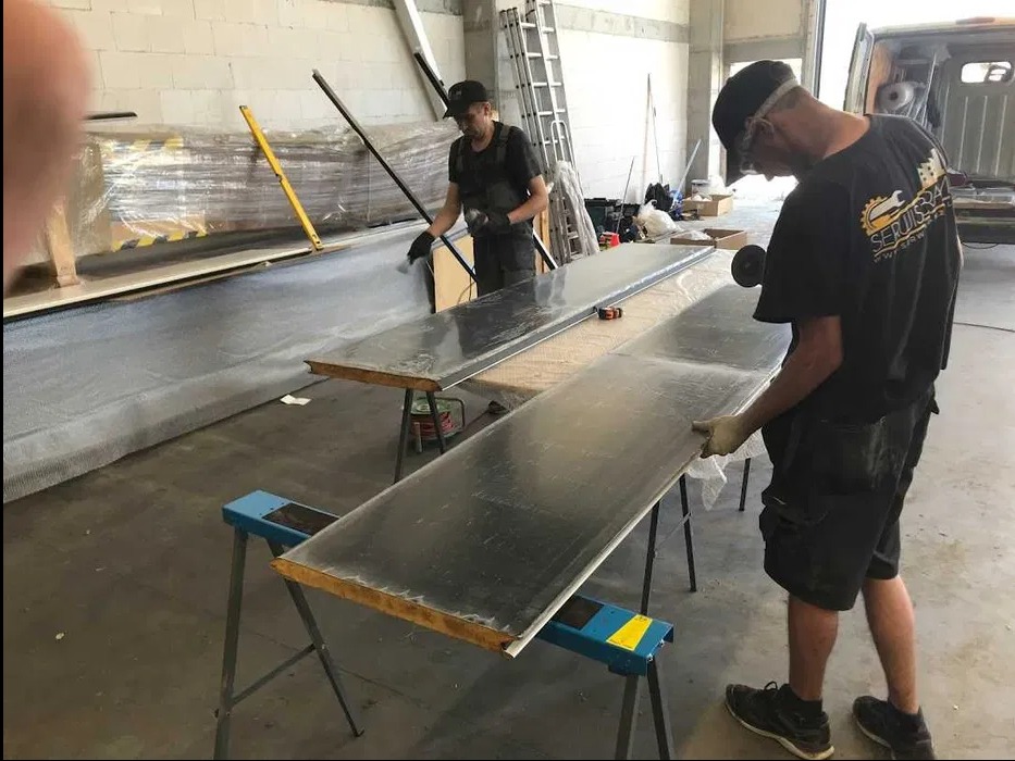 Pracownicy w warsztacie obrabiają metalowe płyty na kozłach, przygotowując elementy do montażu. Widoczne narzędzia i materiały w tle.