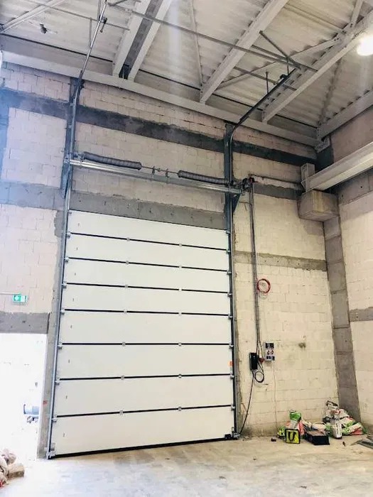 Zamknięta, biała brama garażowa segmentowa z czarnymi liniami, zamontowana w surowym wnętrzu budynku. Widoczne elementy konstrukcyjne i instalacje.