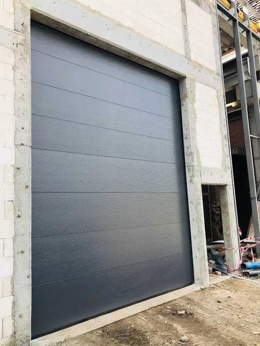 Zamknięta, segmentowa brama garażowa w kolorze antracytu, zamontowana w surowym otworze budynku w trakcie budowy. Widoczne bloczki i stalowa konstrukcja.