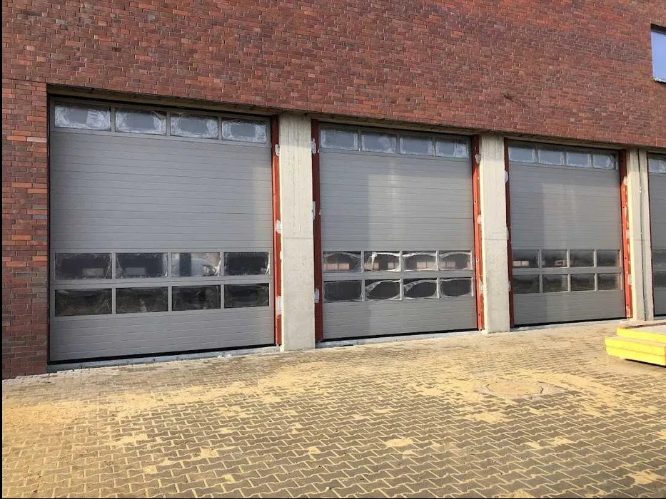 Szara brama przemysłowa segmentowa z oknami, zamontowana w budynku z cegły. Widoczne zabezpieczenia szyb folią. Brama na zewnątrz budynku.