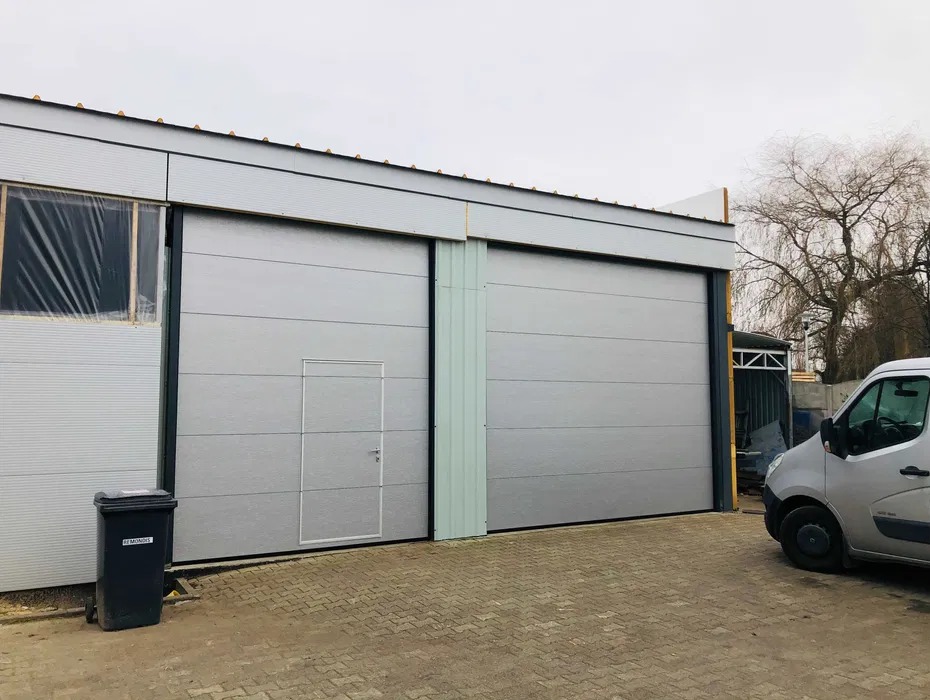 Szara brama przemysłowa z drzwiami w Tychach, widoczna z zewnątrz budynku. Obok zaparkowany srebrny bus i czarny kosz na śmieci.