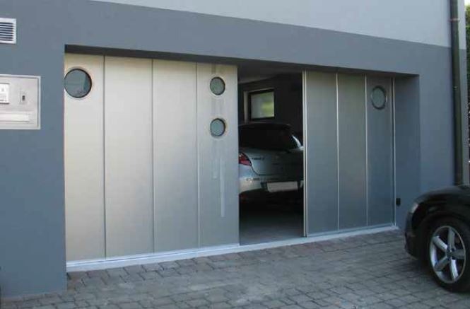 Nowoczesna, srebrna brama garażowa segmentowa z okrągłymi okienkami, częściowo otwarta, ukazująca wnętrze garażu z zaparkowanym samochodem. Elewacja budynku w odcieniach szarości.