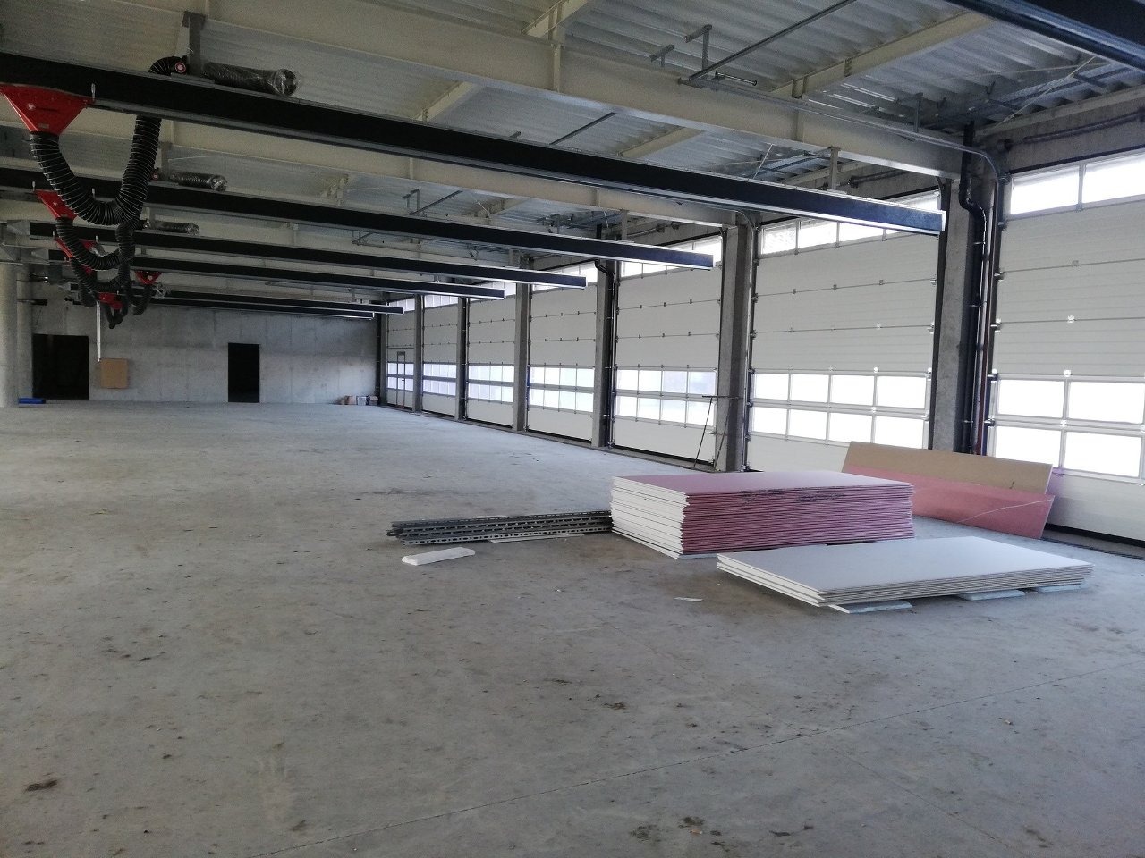Wnętrze hali z segmentowymi bramami garażowymi, widoczne płyty gipsowe i elementy konstrukcyjne sufitu. Surowy beton i stal tworzą industrialny charakter przestrzeni.