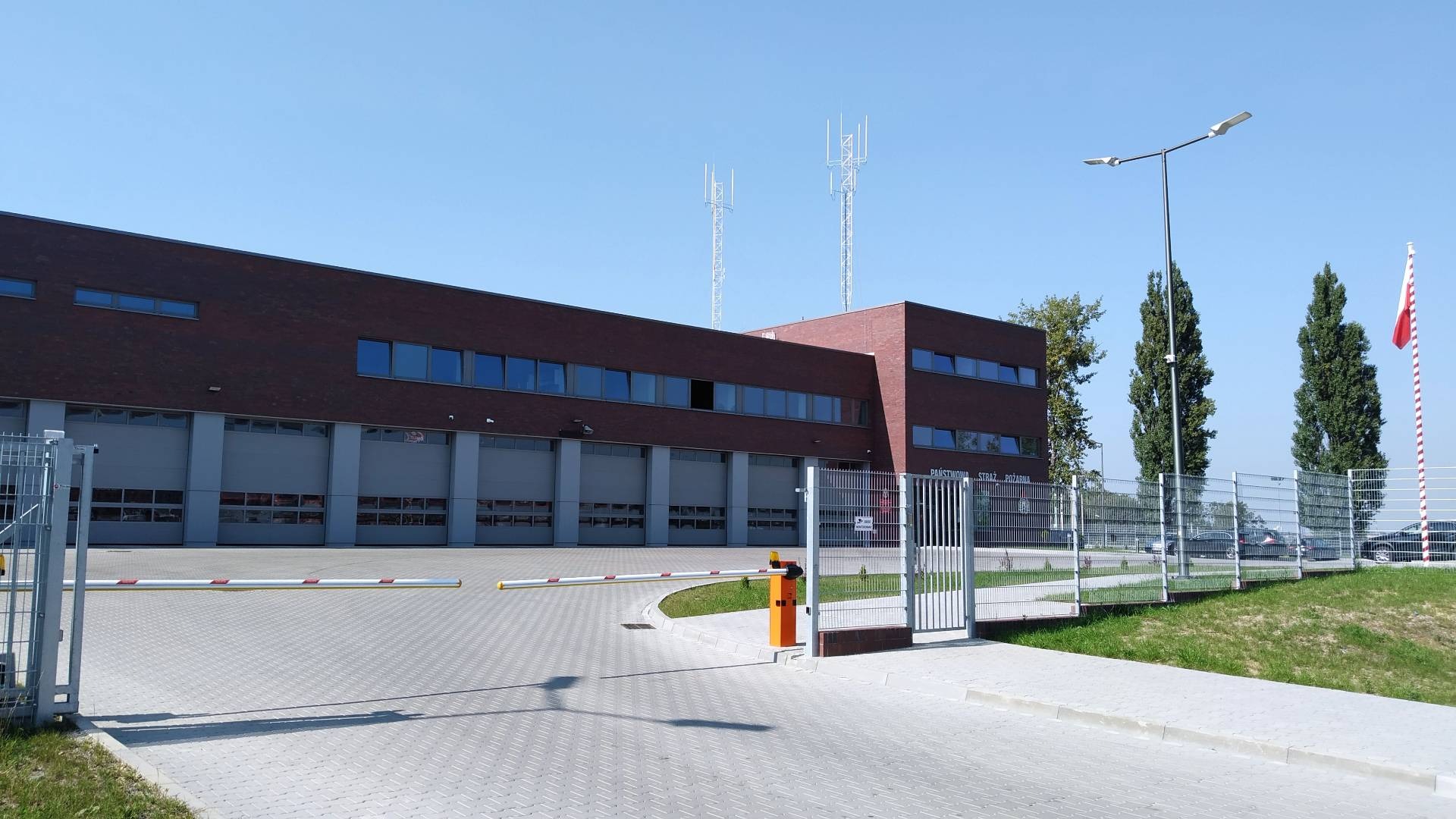 Budynek straży pożarnej z bramami garażowymi, szlabanem i ogrodzeniem z siatki. Widoczne maszty antenowe i flaga Polski. Słoneczny dzień.