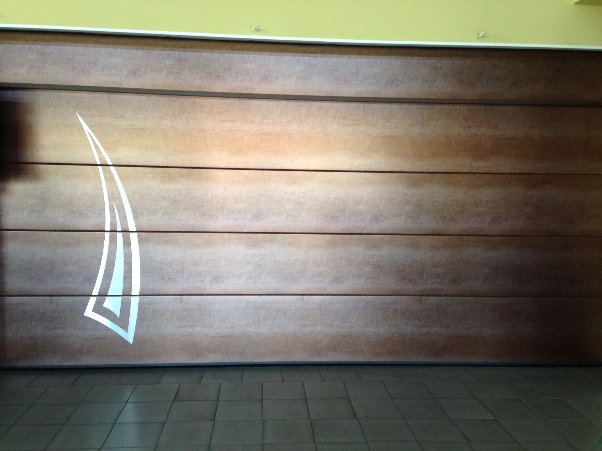 Panelowa brama garażowa w odcieniach brązu z dekoracyjnym, białym motywem graficznym po lewej stronie, widoczna na tle szarej podłogi z płytek.