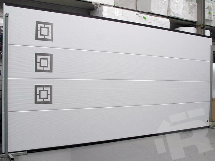 Biała segmentowa brama garażowa z trzema kwadratowymi zdobieniami, widoczna z boku, w magazynie. Minimalistyczny design z subtelną teksturą paneli.
