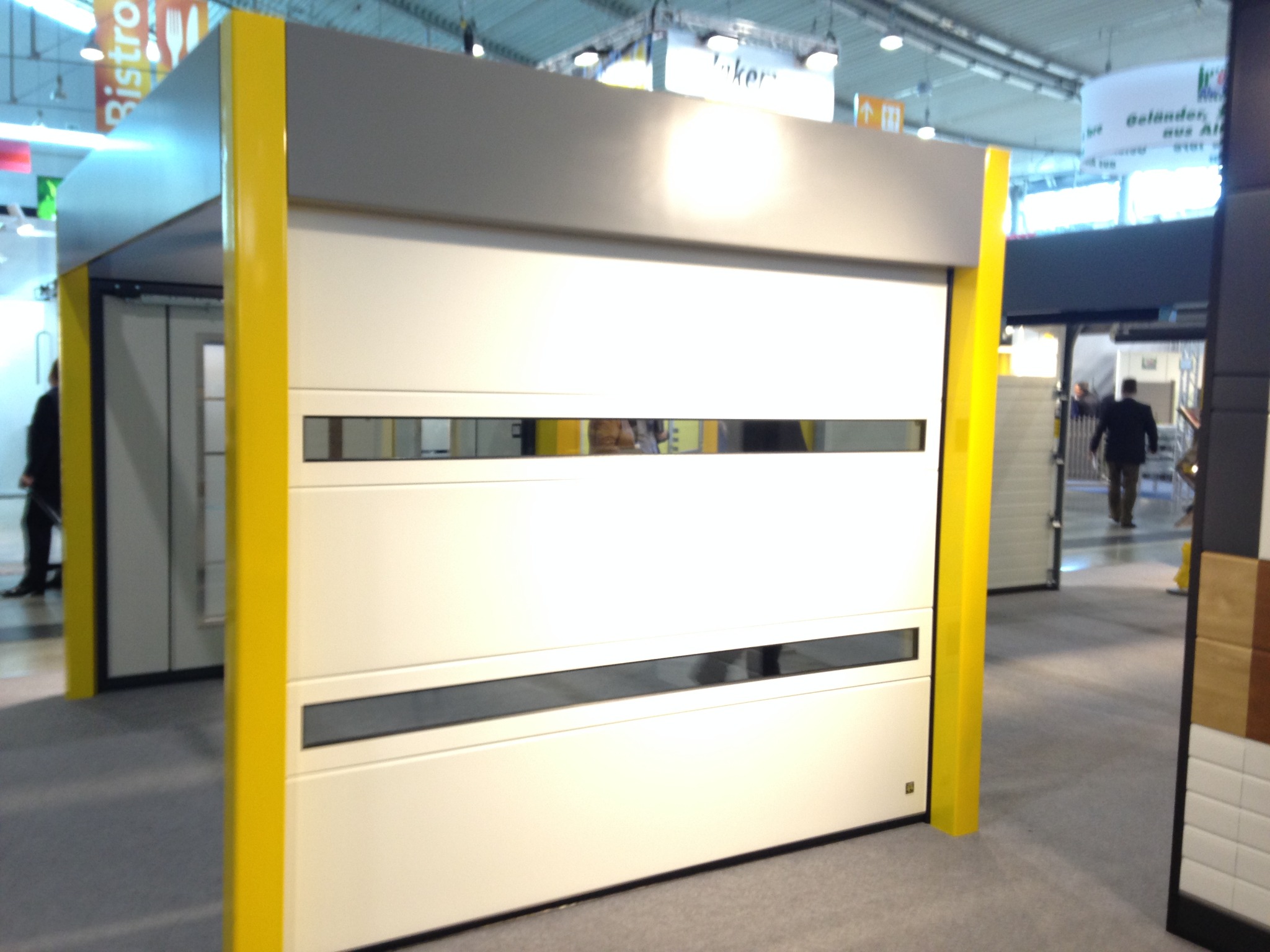 Biała brama garażowa segmentowa z poziomymi przeszkleniami, obramowana jaskrawożółtą ramą. Prezentacja na targach, widoczne inne ekspozycje w tle.