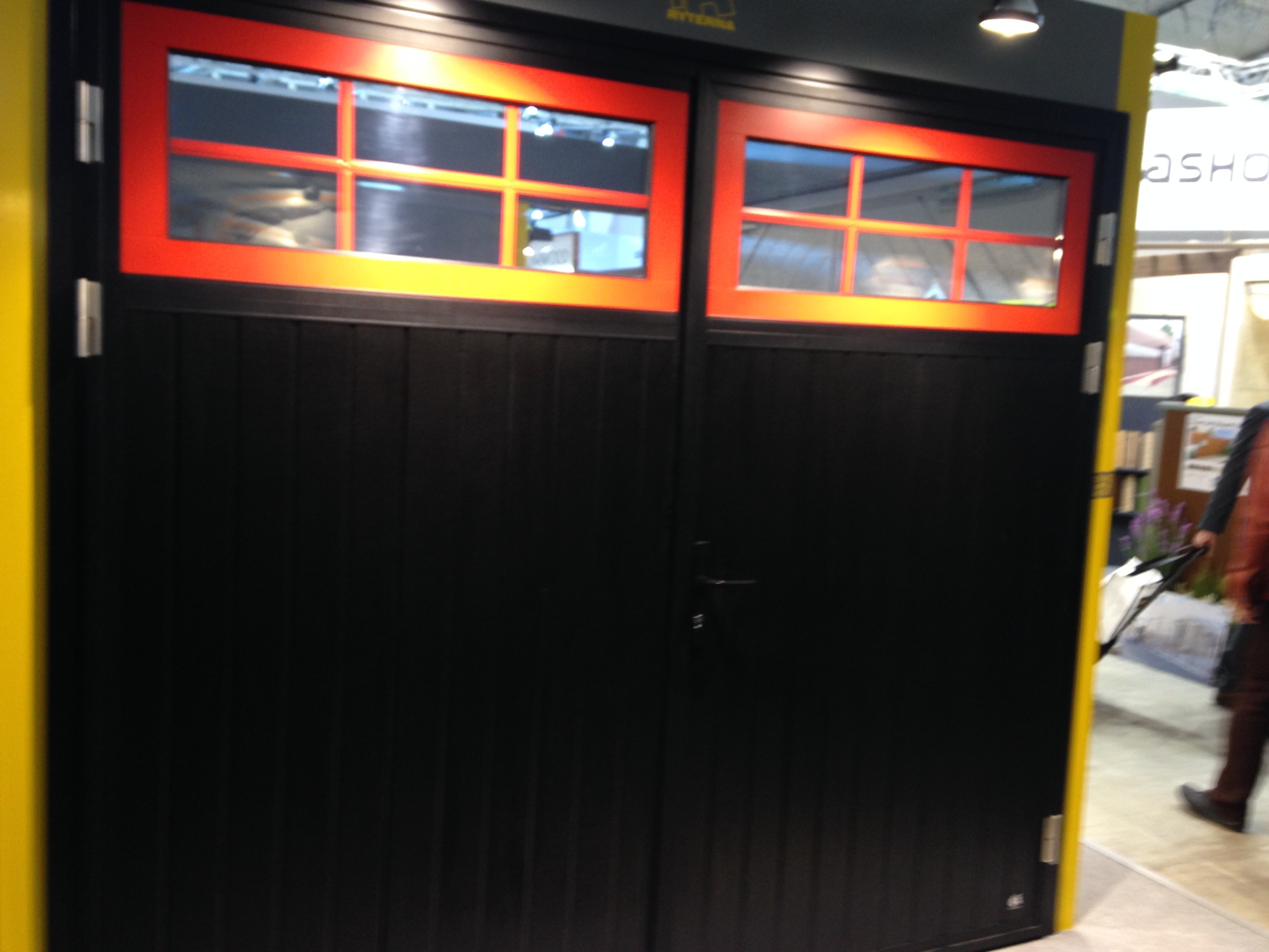 Czarna brama garażowa z czerwonymi, przeszklonymi panelami w górnej części, widoczna na tle żółtej ściany. Brama dwuskrzydłowa z pionowymi panelami.