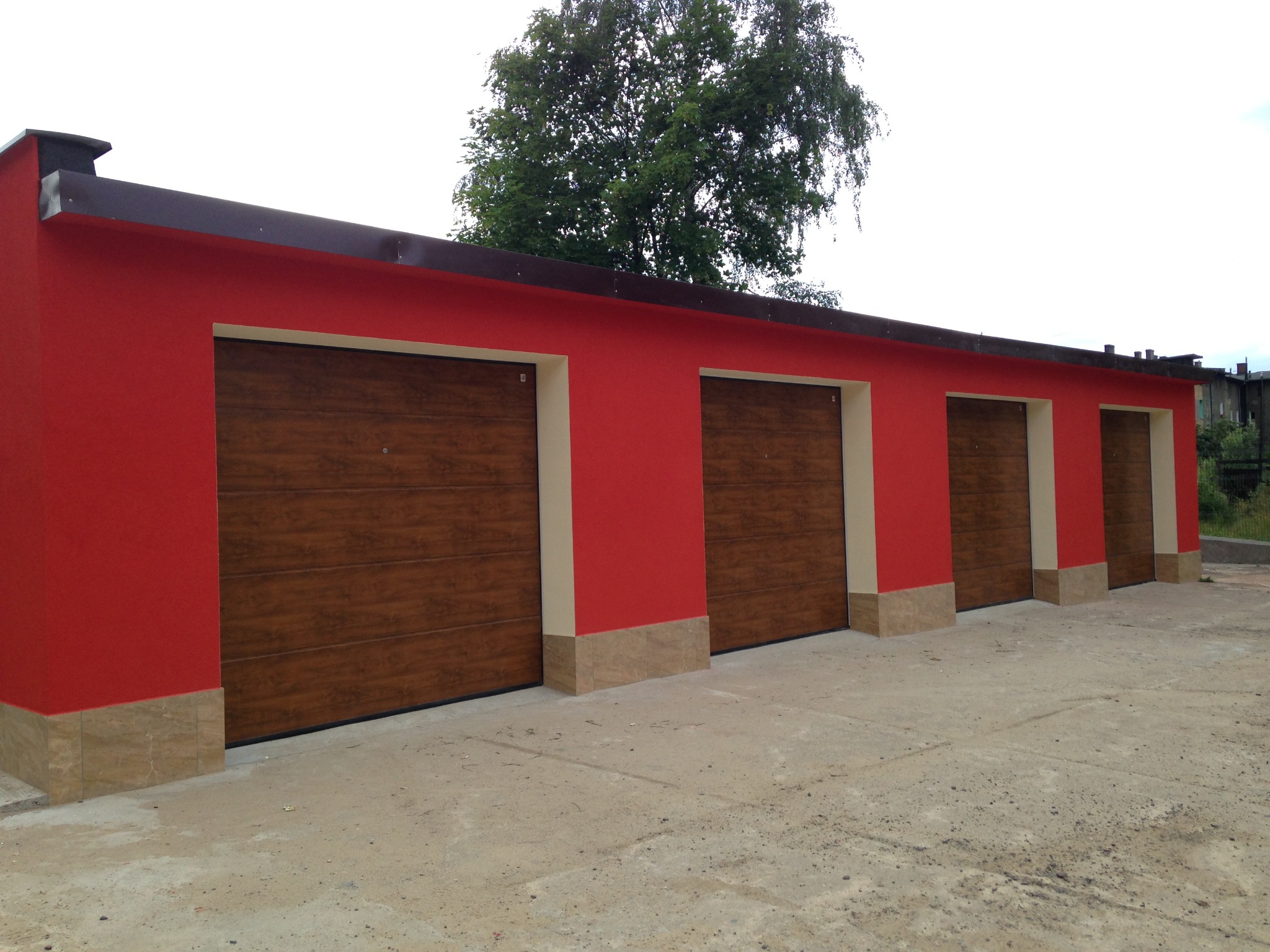 Czerwony budynek garażowy z czterema brązowymi bramami segmentowymi w Katowicach. Widok z zewnątrz, jasne tło, drzewo w tle.
