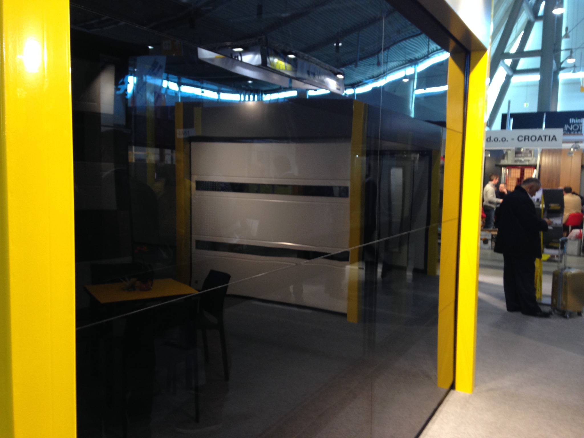Biała segmentowa brama garażowa z czarnymi akcentami, widoczna przez szklaną ścianę z żółtą ramą, na tle hali wystawienniczej z ludźmi.