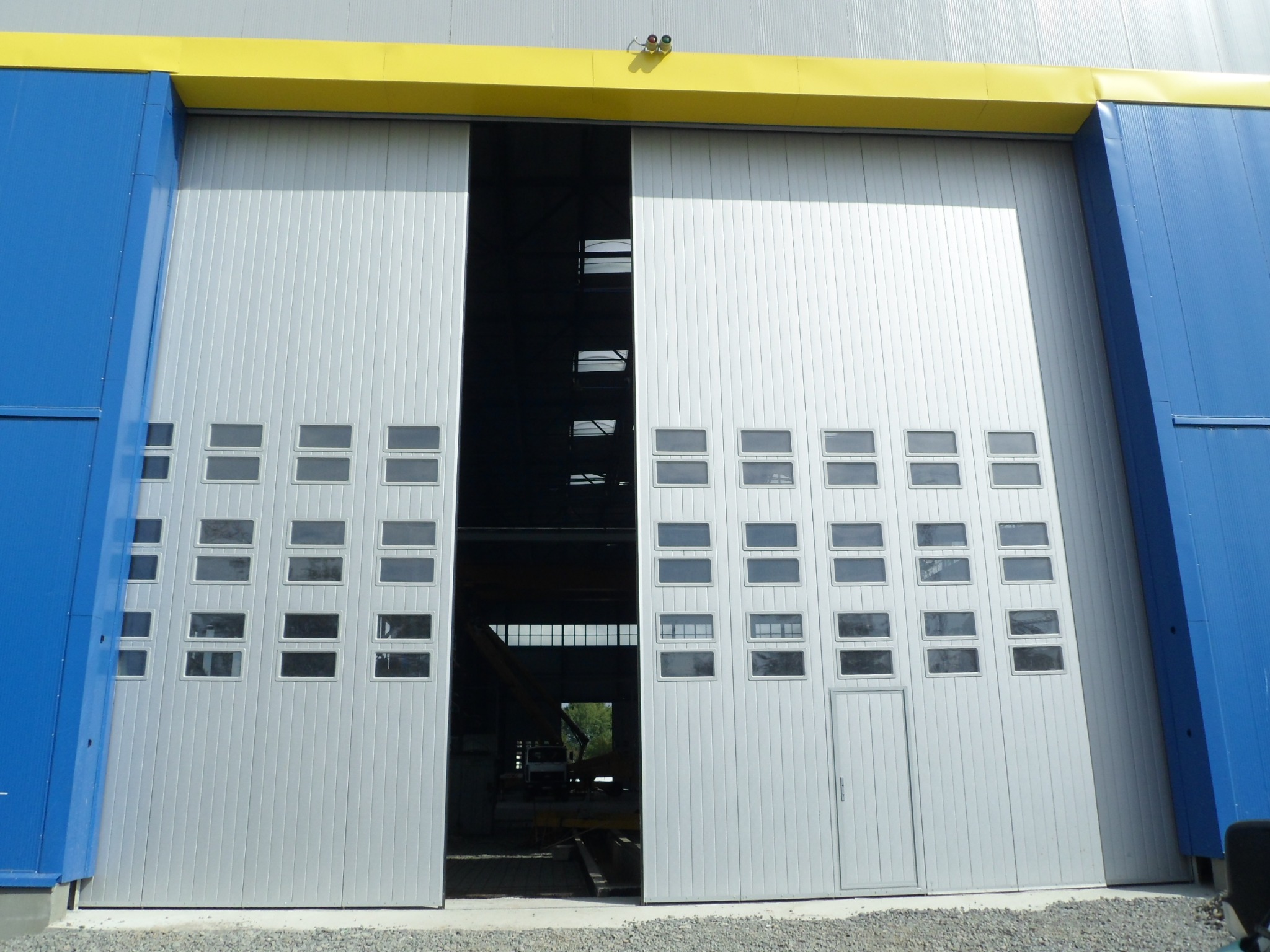 Otwarta, przesuwna brama przemysłowa z oknami i drzwiami, w kolorze srebrnym, z niebieskimi i żółtymi elementami elewacji budynku. Widok na wnętrze hali.