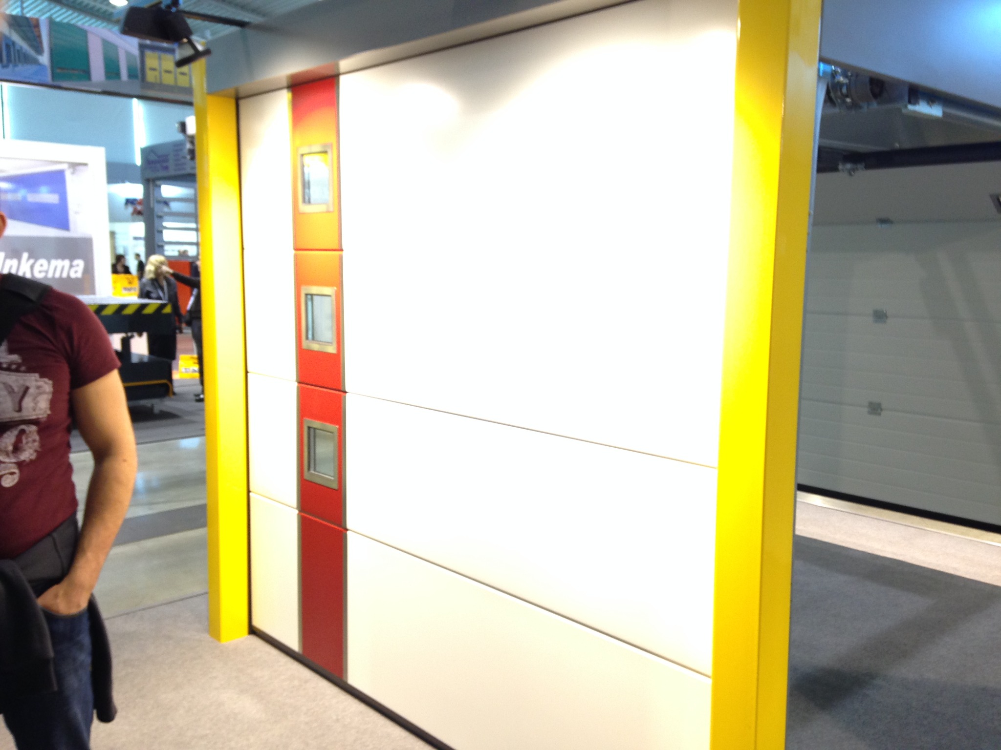 Biała brama garażowa segmentowa z trzema małymi okienkami w czerwonym pasie, obramowana żółtą ramą, prezentowana na targach.