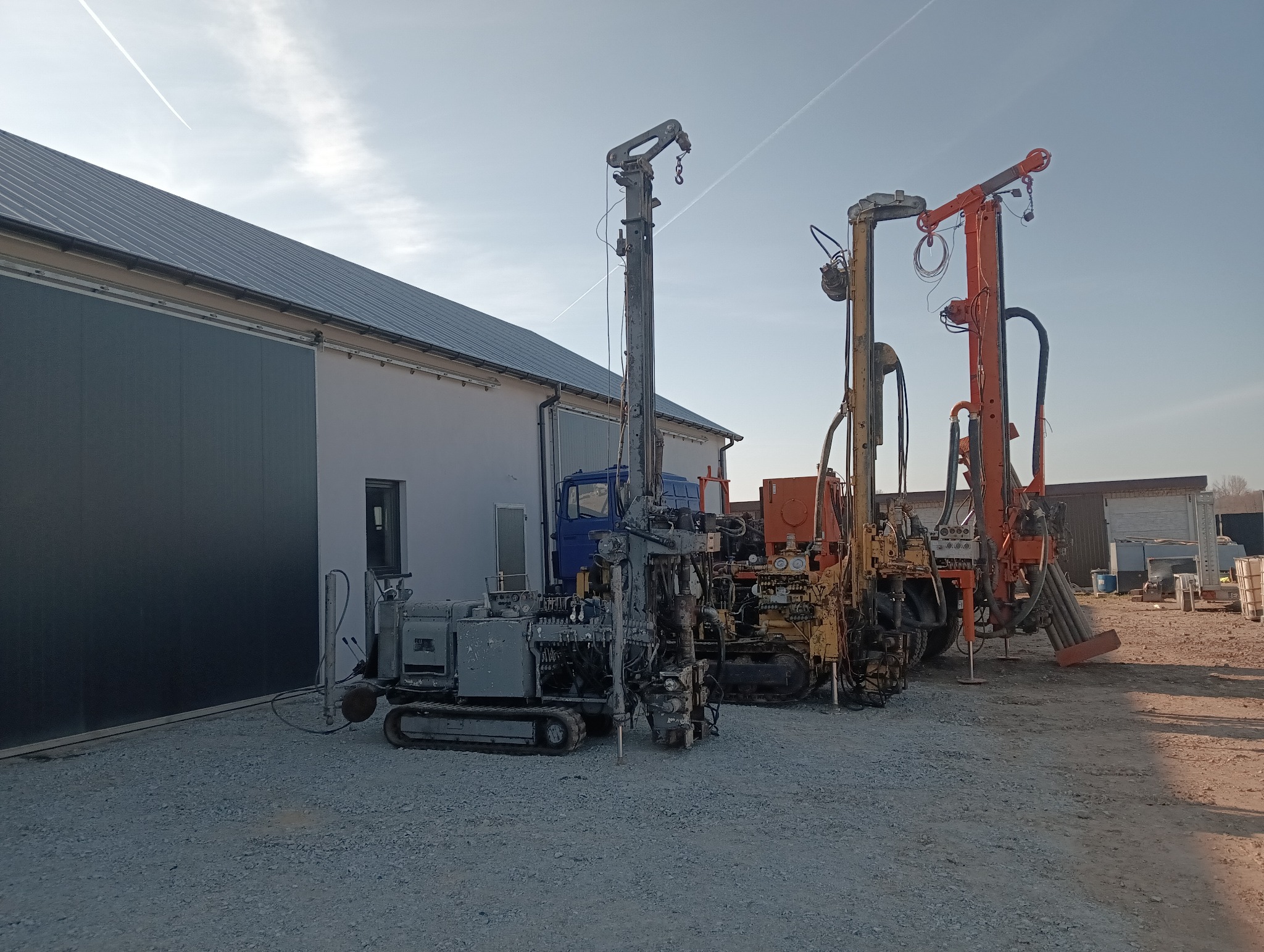 Różne wiertnice ustawione obok budynku, w tym jedna gąsienicowa. W tle widoczny niebieski samochód ciężarowy. Nad nimi smuga kondensacyjna samolotu.