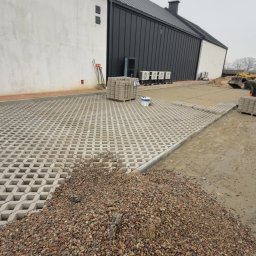 Dawid Dziuba - Układanie ażurowych płyt i wypełnianie ich kamieniem w Kwilcz. Widoczne narzędzia i materiały budowlane. Częściowo ułożona nawierzchnia na tle budynku.