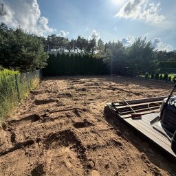 Dawid Dziuba - Przygotowany teren pod budowę tarasu z widocznymi śladami maszyn, ułożonymi deskami i narzędziami. Słoneczny dzień, drzewa w tle.