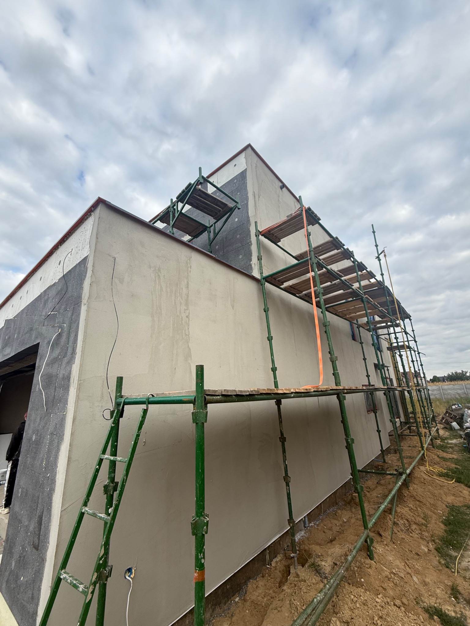 Ściana budynku w trakcie prac wykończeniowych, z nałożonym tynkiem i rusztowaniem. Widoczne ocieplenie i fragment instalacji elektrycznej. Ujęcie z dołu.