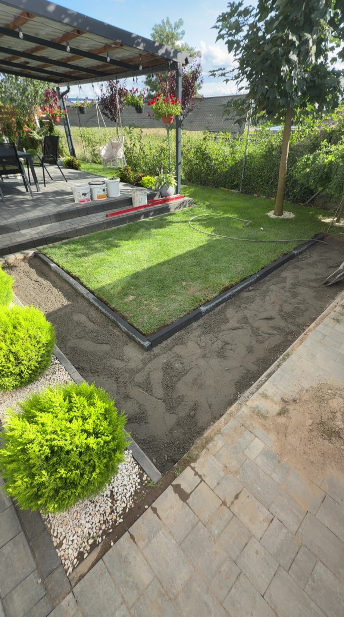 Przygotowanie podłoża pod ścieżkę w ogrodzie: wyrównana ziemia, obrzeża, kostka brukowa w tle, krzewy ozdobne i trawnik. Widok z góry.