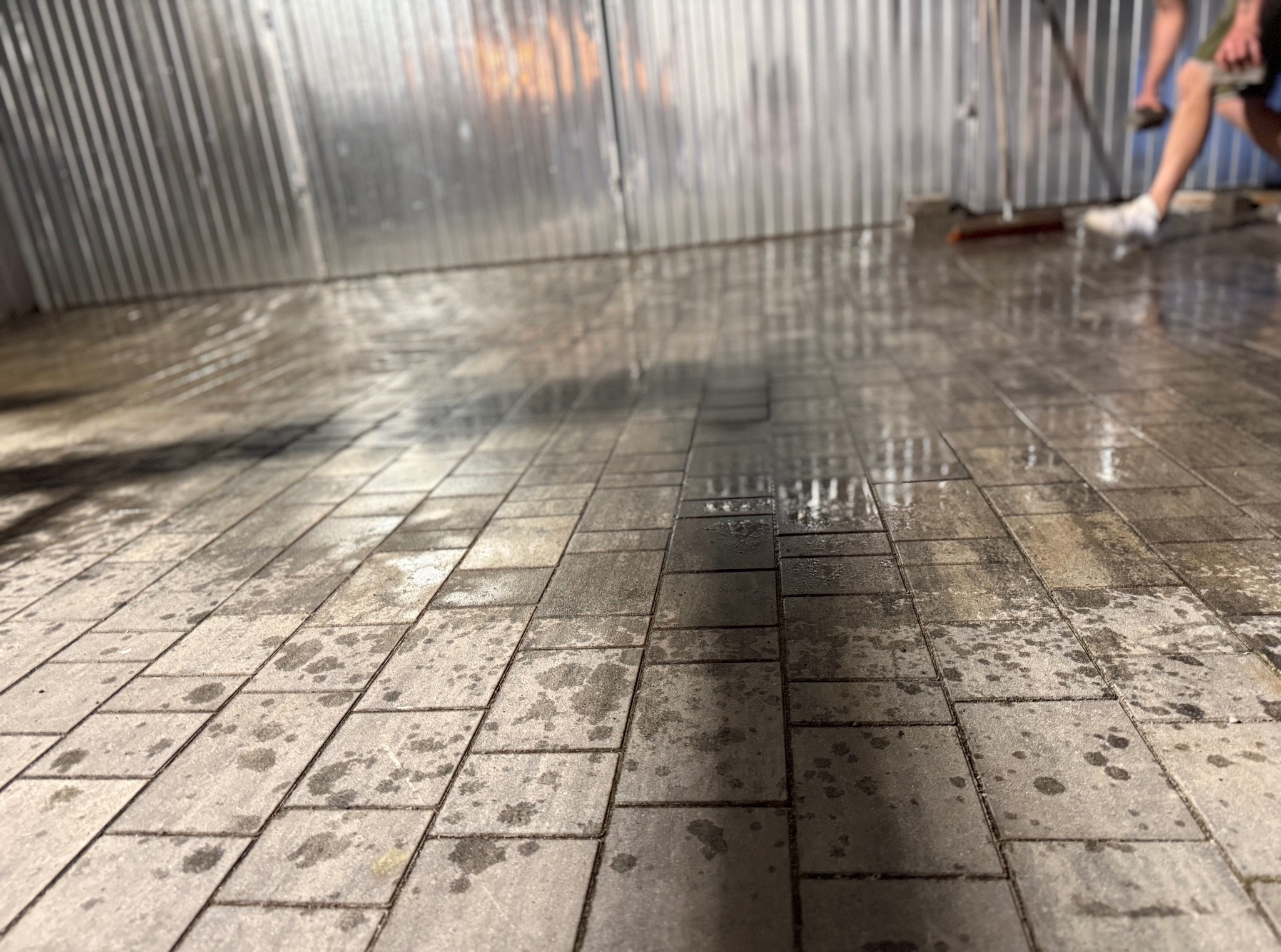 Umyta, szara kostka brukowa pod kątem, z odbiciami. W tle blaszana ściana i fragment osoby z miotłą. Ślady wody na kostce podkreślają jej strukturę.