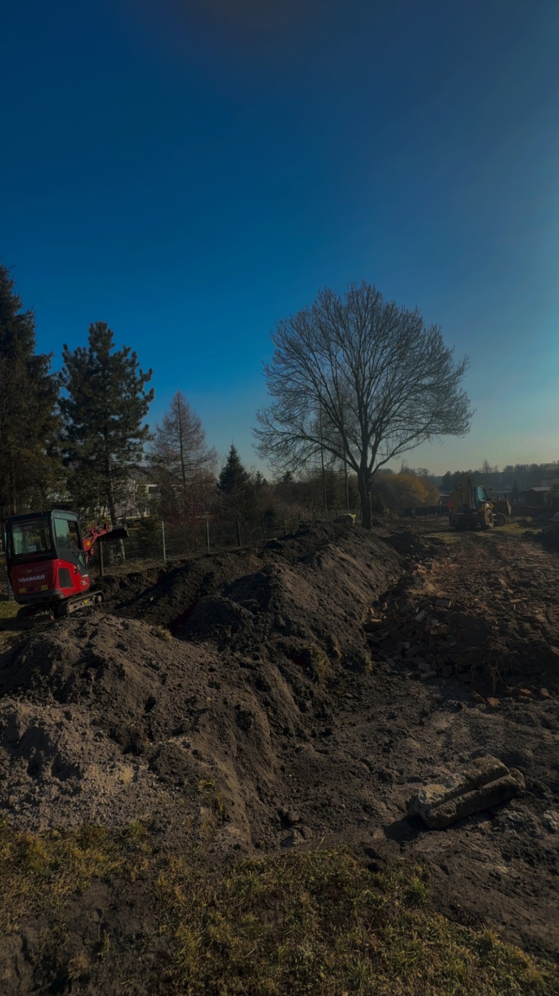 Czerwona mini koparka Yanmar przy wykopie ziemi na tle drzew i błękitnego nieba. W oddali widoczna większa koparka. Prace ziemne w toku.