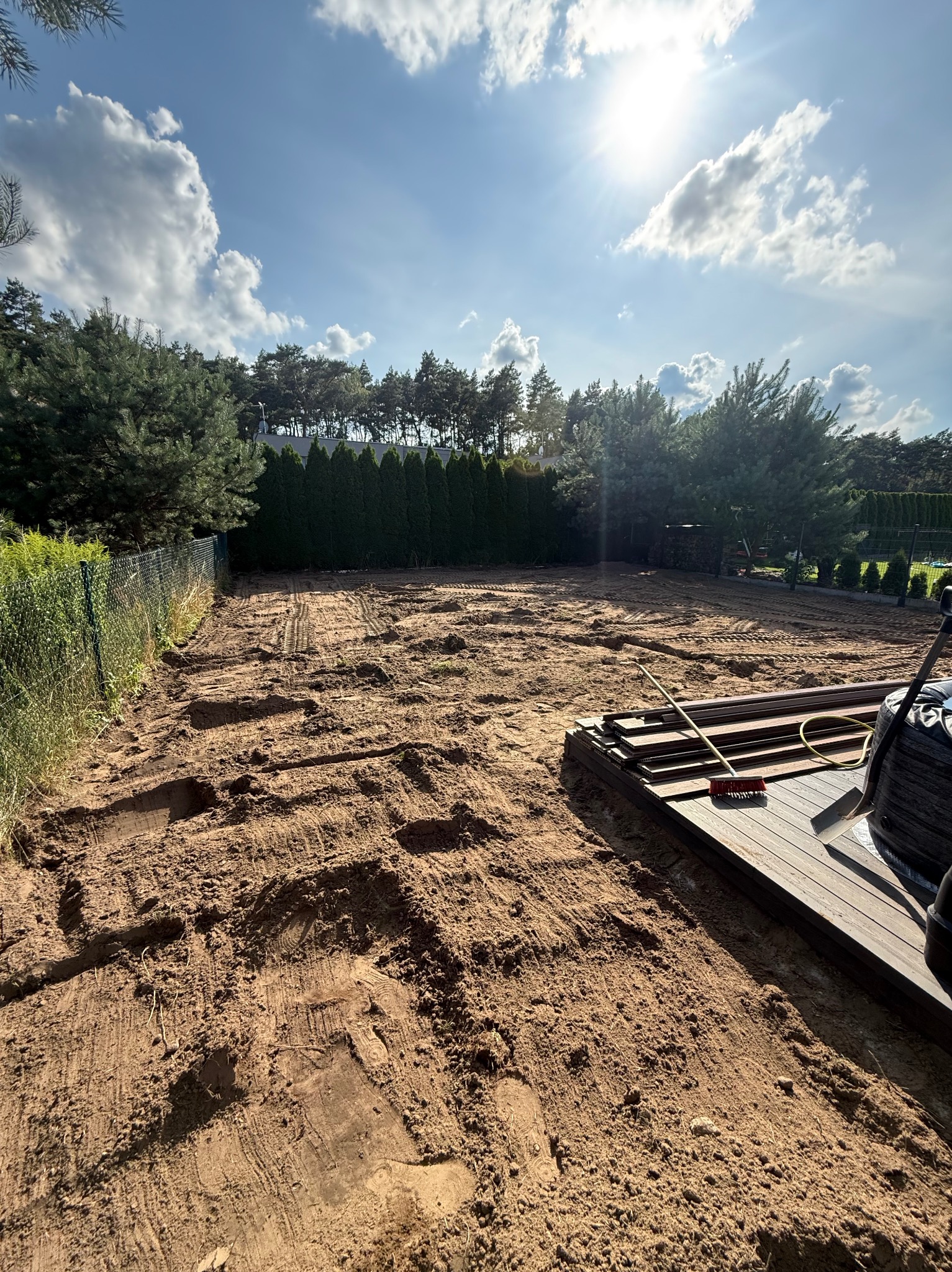 Przygotowany teren pod budowę tarasu z widocznymi śladami maszyn, ułożonymi deskami i narzędziami. Słoneczny dzień, drzewa w tle.