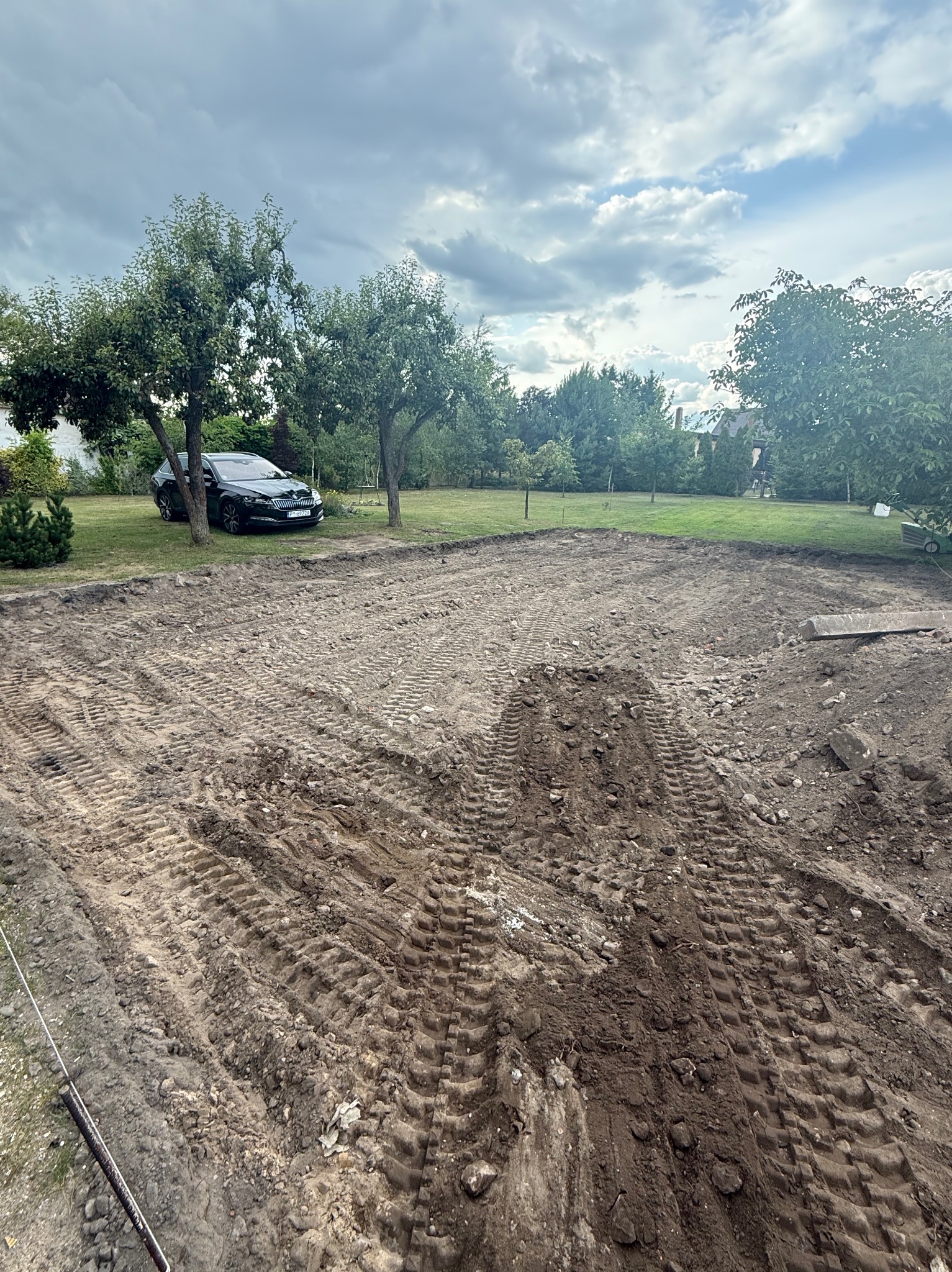 Przygotowany teren pod budowę z widocznymi śladami opon, w tle drzewa i czarny samochód osobowy. Pochmurne niebo.