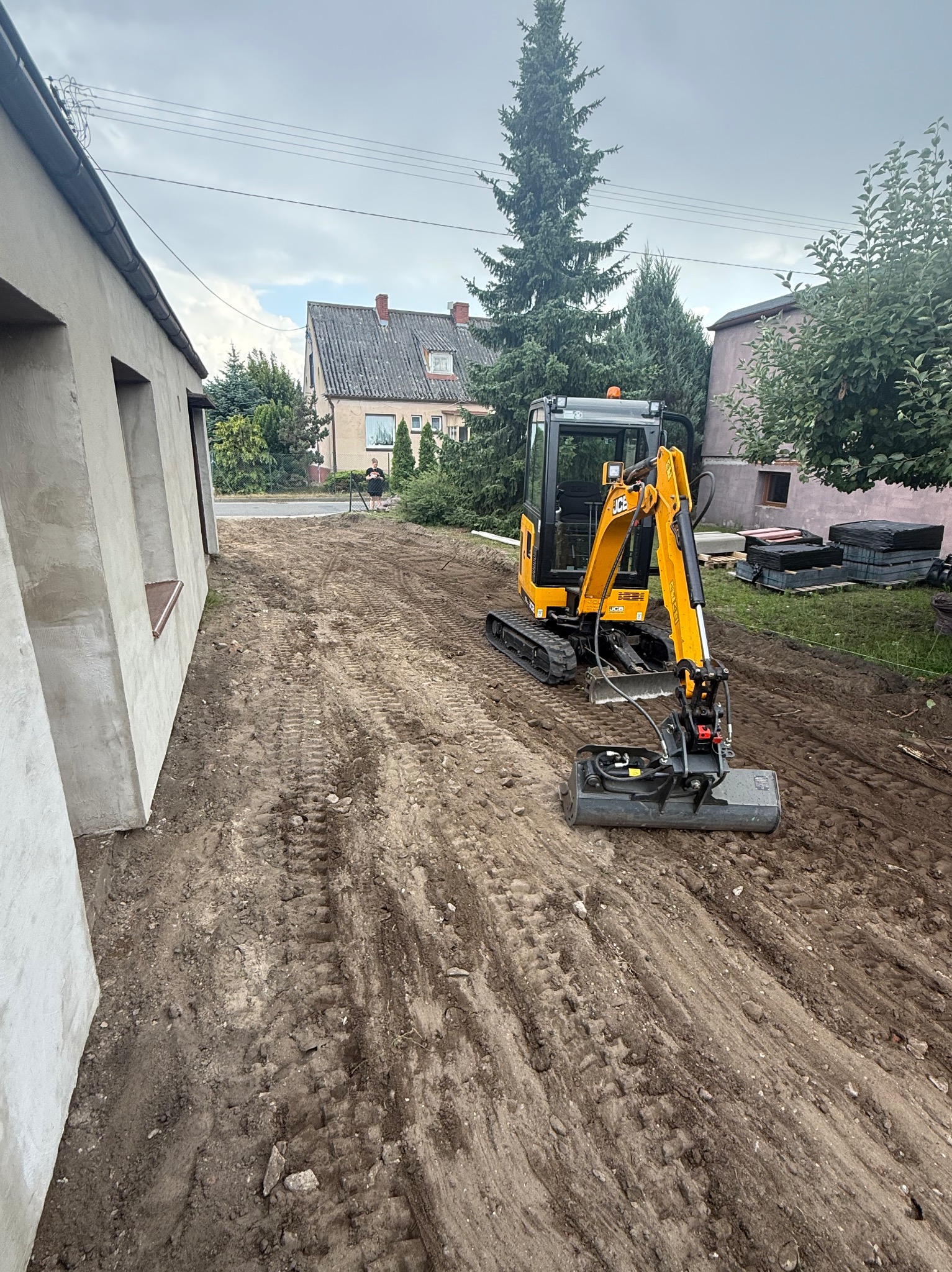 Żółta minikoparka JCB z frezarką do pni na nierównym, przygotowanym terenie budowy. W tle budynki mieszkalne i drzewa. Pochmurne niebo.