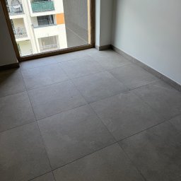 Tara Den - Wykończony pokój z szarymi płytkami podłogowymi i dużym oknem z widokiem na balkon innego budynku. Minimalistyczny design, neutralna kolorystyka.