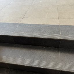 Tara Den - Wykończone schody z płytek w odcieniach szarości i beżu, z detalami antypoślizgowymi. Widoczne połączenia między płytkami i fragment ściany w tle.