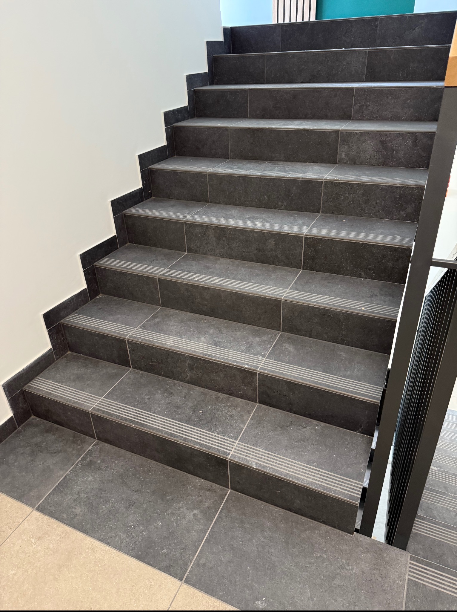 Schody wyłożone ciemnoszarą, matową płytką z antypoślizgowymi paskami. Ujęcie z dołu, widoczny fragment metalowej barierki i jasne ściany. Estetyczne wykończenie.