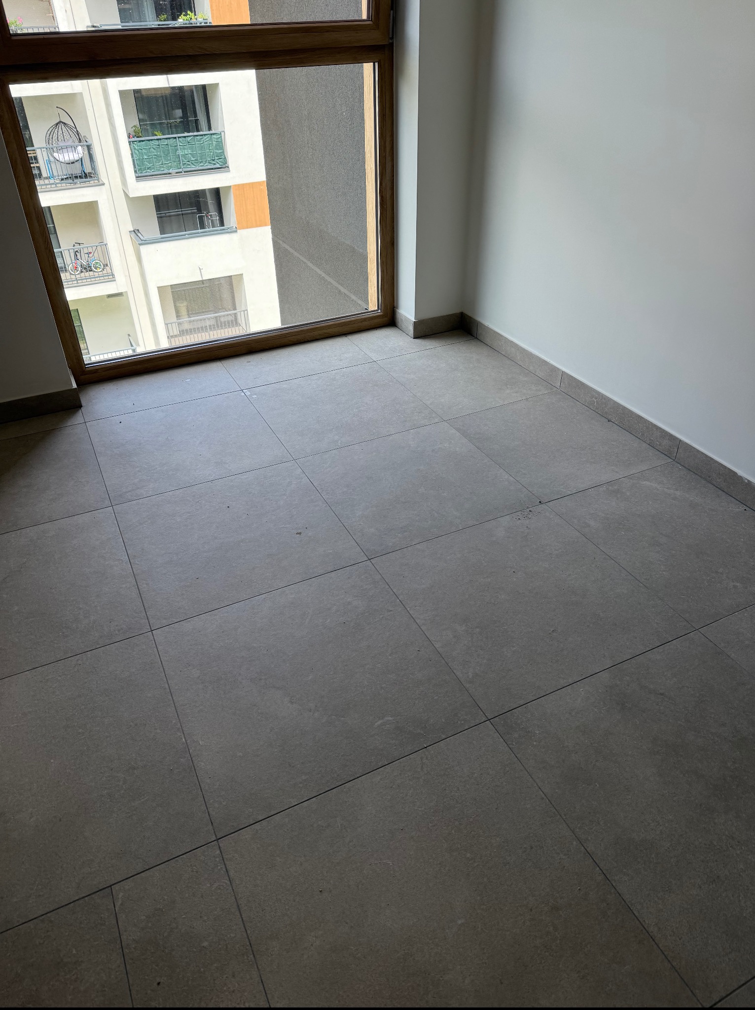 Wykończony pokój z szarymi płytkami podłogowymi i dużym oknem z widokiem na balkon innego budynku. Minimalistyczny design, neutralna kolorystyka.