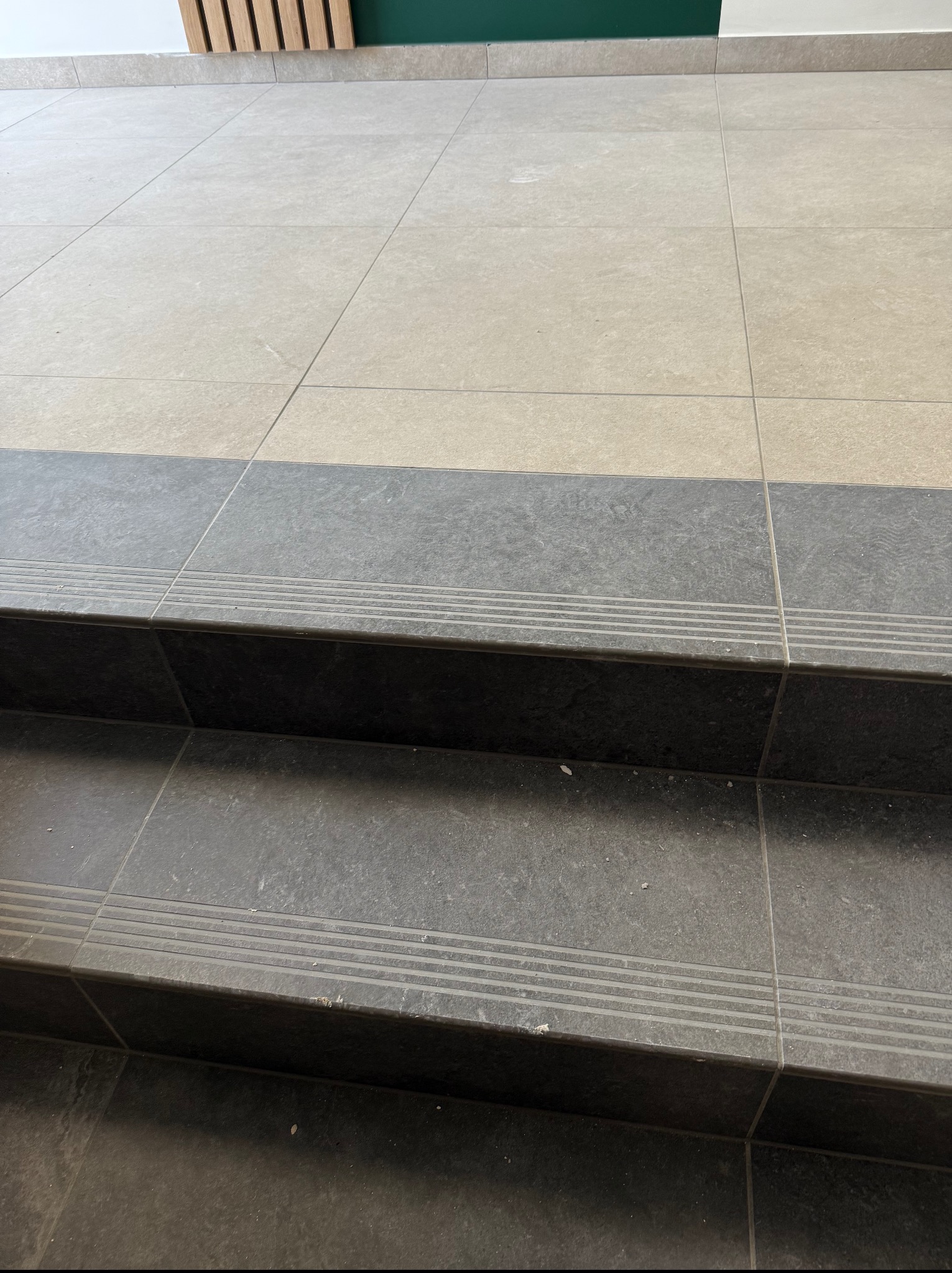 Wykończone schody z płytek w odcieniach szarości i beżu, z detalami antypoślizgowymi. Widoczne połączenia między płytkami i fragment ściany w tle.