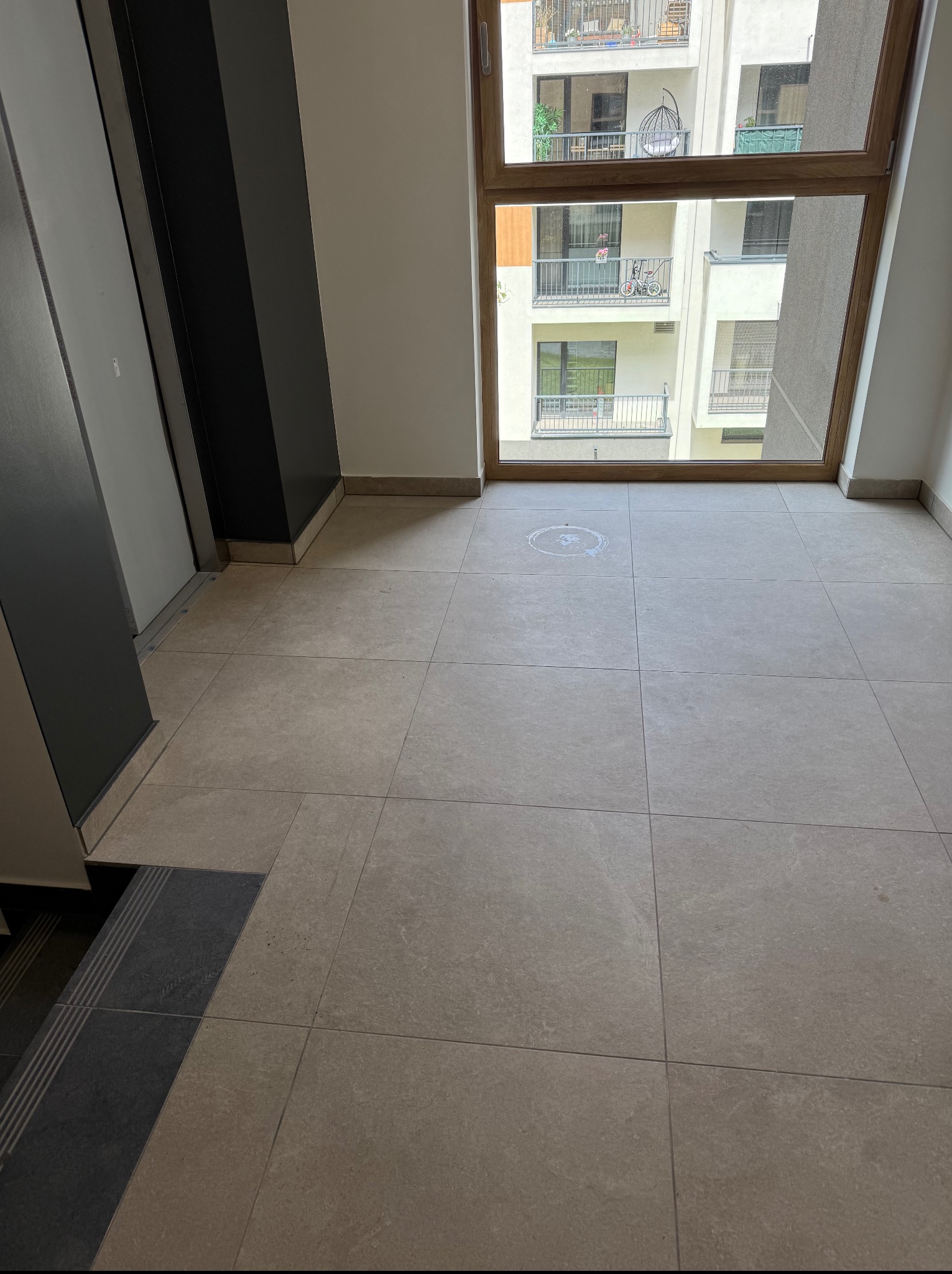 Beżowe płytki podłogowe w korytarzu apartamentowca, z widokiem na balkony przez okno. Ciemne schody i fragment windy po lewej stronie kadru.