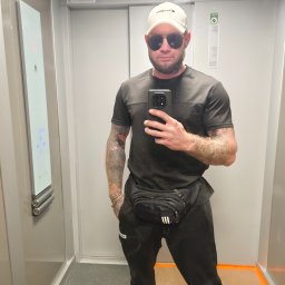 Anton Suvorov - Mężczyzna w windzie robi selfie telefonem. Widoczne tatuaże na rękach, czarne ubranie, biała czapka i okulary przeciwsłoneczne. W tle panel sterowania windą.