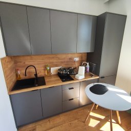 Anton Suvorov - Szare szafki kuchenne z drewnianym blatem, czarny zlew i płyta grzewcza, biały stół z czarną paterą, zielona zasłona w tle. Widok z góry na małą kuchnię.