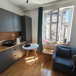 Anton Suvorov - Jasne wnętrze mieszkania z szarymi meblami kuchennymi i drewnianym blatem, okno z widokiem na budynek, drewniana podłoga, mały stolik i sofa.