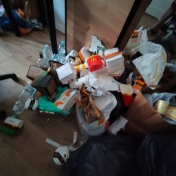 Anton Suvorov - Stos śmieci, w tym pudełka i butelki po napojach, leżący na podłodze w rogu pokoju, blisko nogi stołu. Widoczny czarny worek na śmieci.