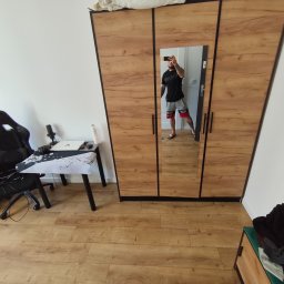 Anton Suvorov - Pokój z drewnianą podłogą i szafą z lustrem, w którym odbija się mężczyzna robiący zdjęcie telefonem. Widoczne biurko z krzesłem gamingowym i uchwyt na telewizor na ścianie.