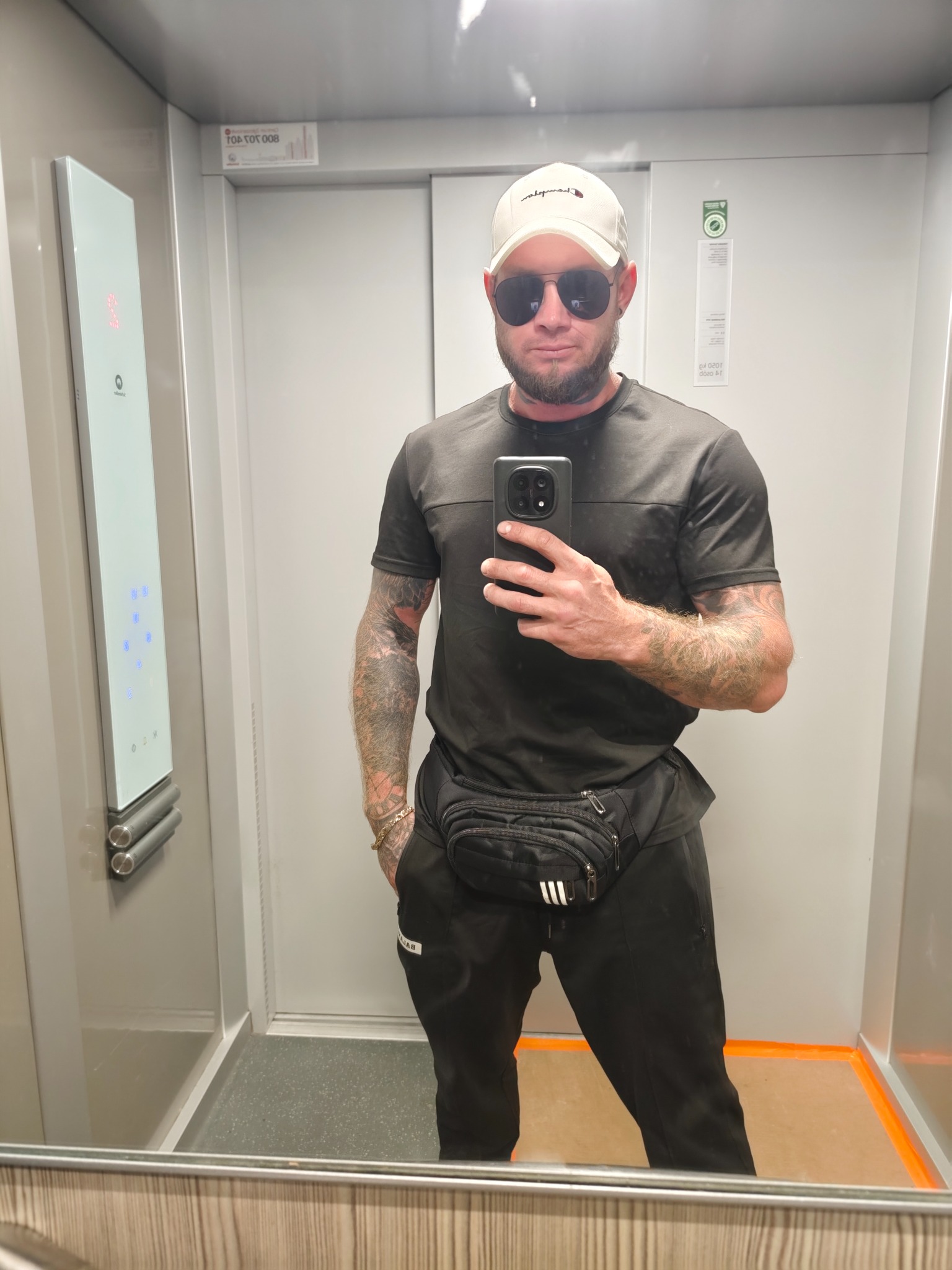 Mężczyzna w windzie robi selfie telefonem. Widoczne tatuaże na rękach, czarne ubranie, biała czapka i okulary przeciwsłoneczne. W tle panel sterowania windą.