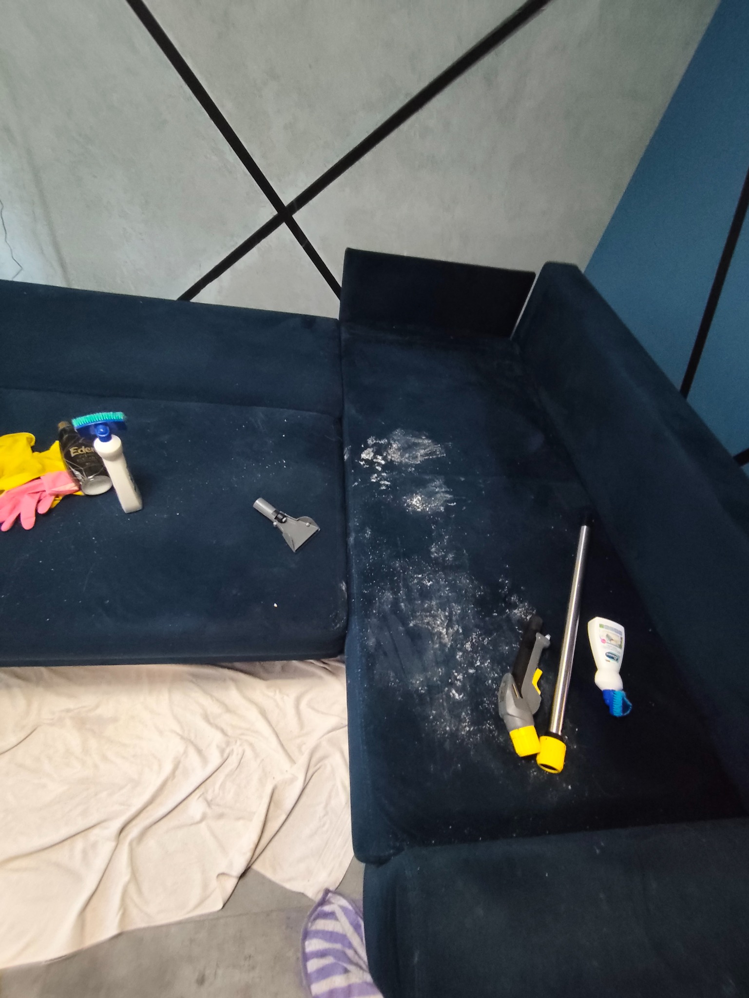 Ciemnoniebieska sofa narożna w trakcie czyszczenia. Widoczne środki czyszczące, końcówki do prania tapicerki i obszar z widocznymi plamami przed usunięciem.