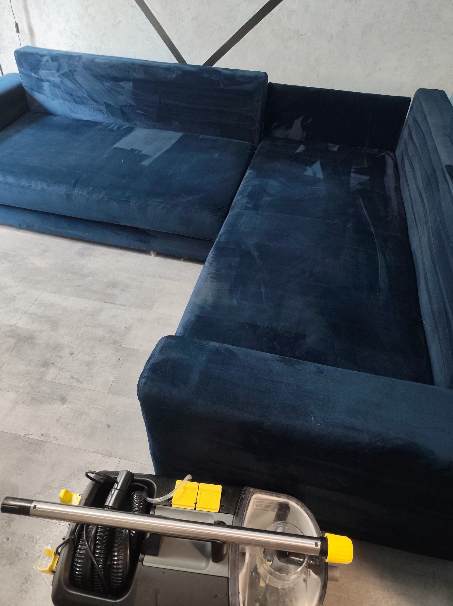 Granatowa, welurowa kanapa narożna po praniu, z widocznymi śladami wilgoci. Na pierwszym planie fragment odkurzacza piorącego z elementami w kolorze żółtym.