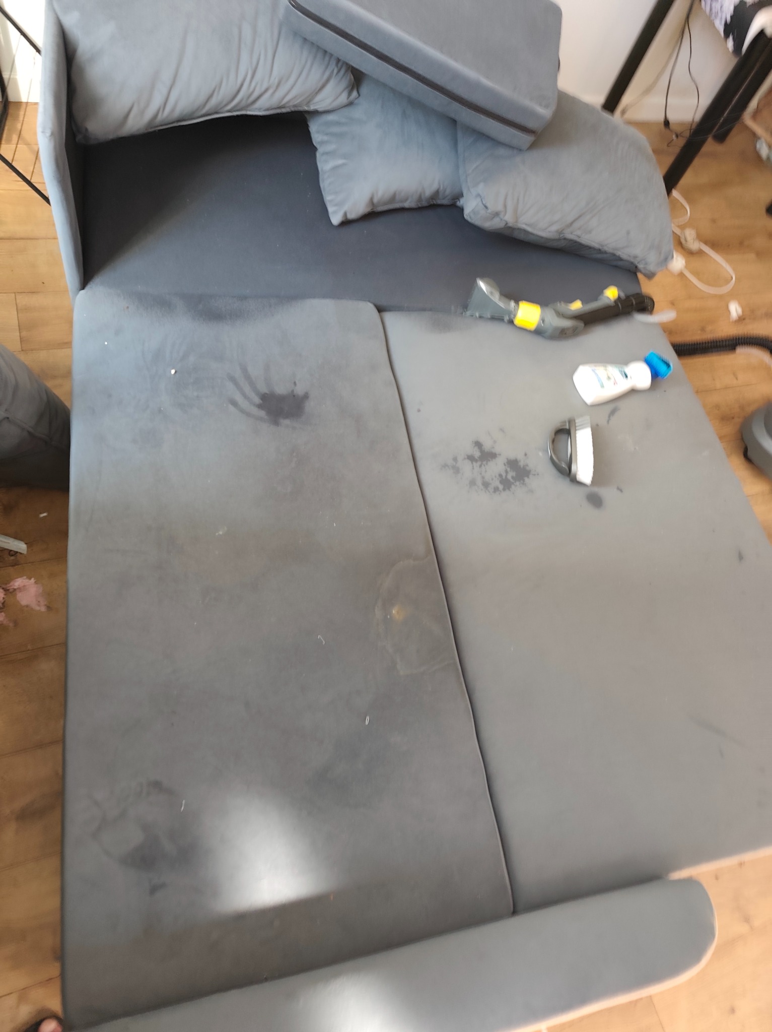 Szara rozkładana sofa z widocznymi plamami i akcesoriami do czyszczenia tapicerki, takimi jak szczotka i odkurzacz piorący. Widoczne zabrudzenia na siedzisku.