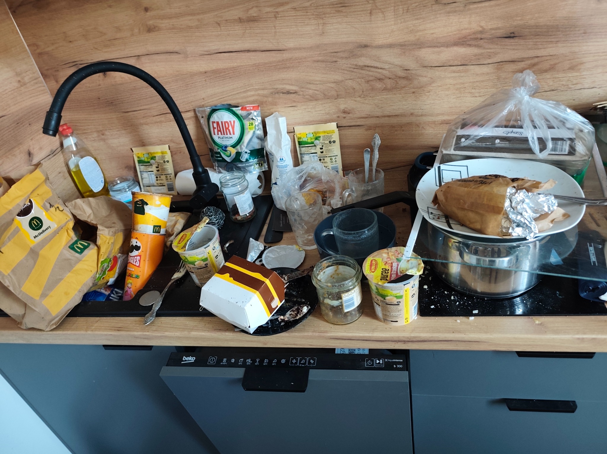 Zabrudzony blat kuchenny z resztkami jedzenia, opakowaniami po posiłkach, brudnymi naczyniami i środkami czystości. Widoczna zmywarka Beko.