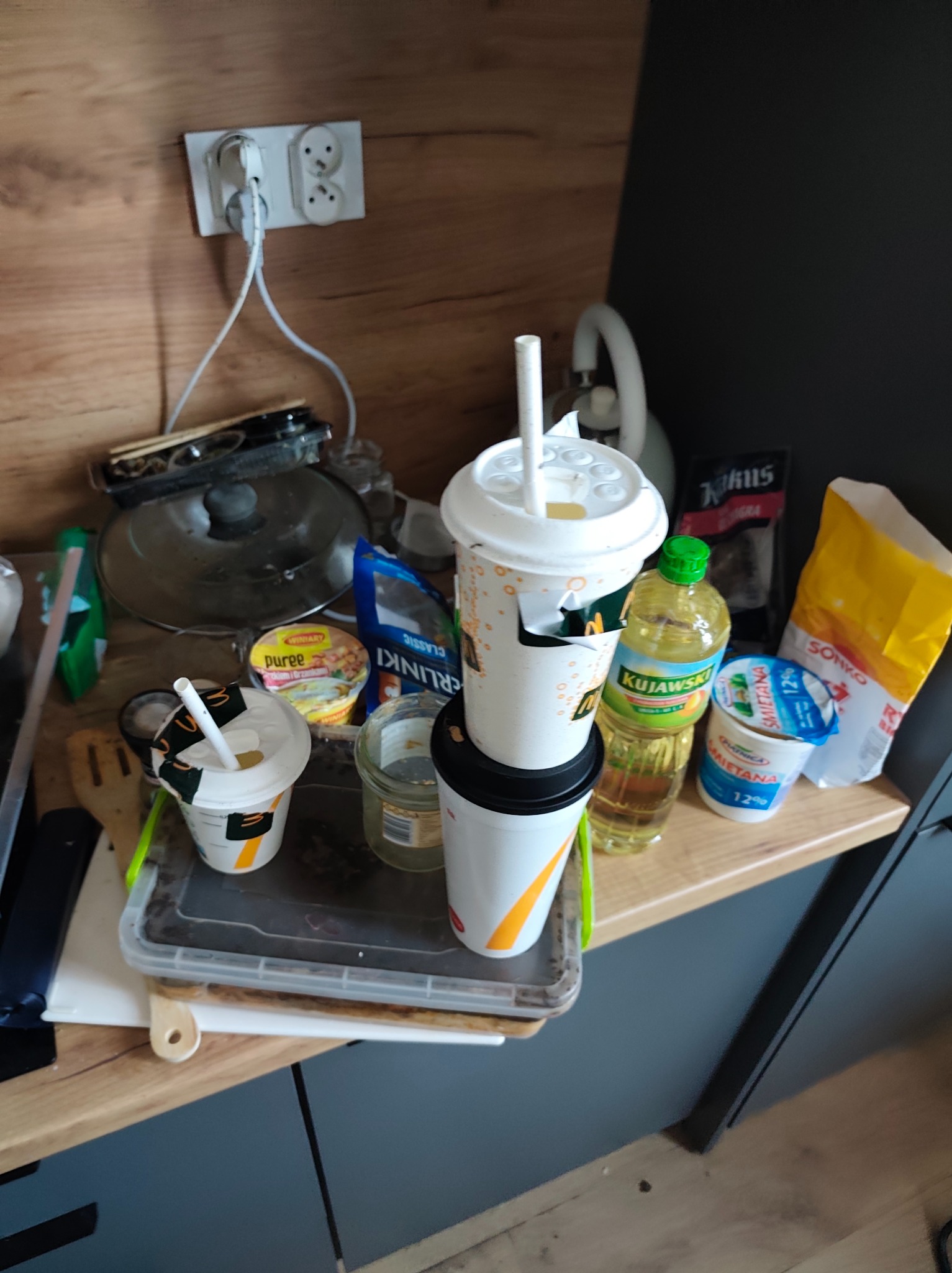 Niezorganizowany blat kuchenny z resztkami jedzenia, kubkami po napojach, olejem Kujawski i produktami spożywczymi, ukazujący potrzebę sprzątania i ogarnięcia przestrzeni.