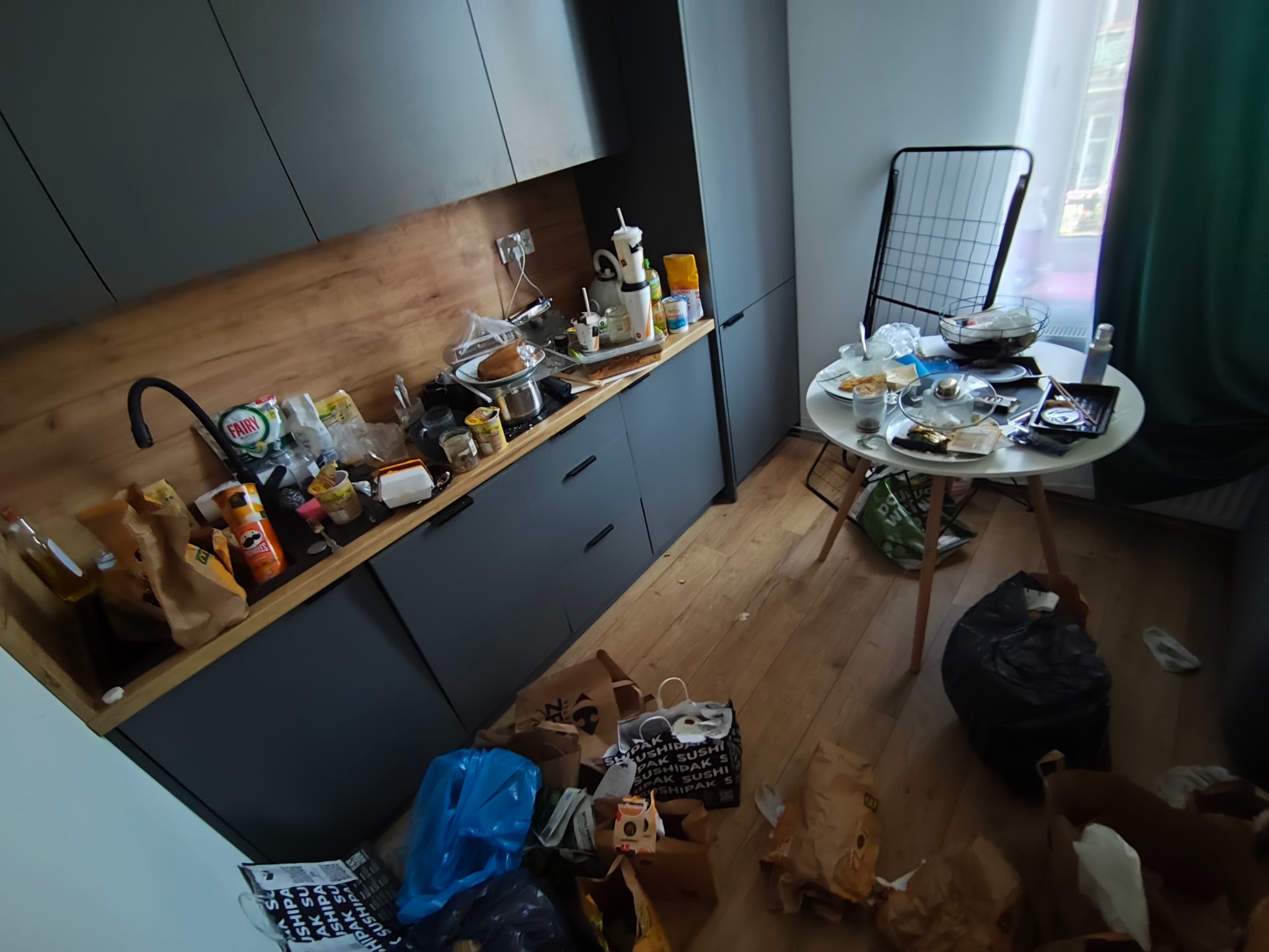 Zabałaganiona kuchnia z niedojedzonym jedzeniem na stole i stercie śmieci na podłodze, widok z góry. Sytuacja przed sprzątaniem.