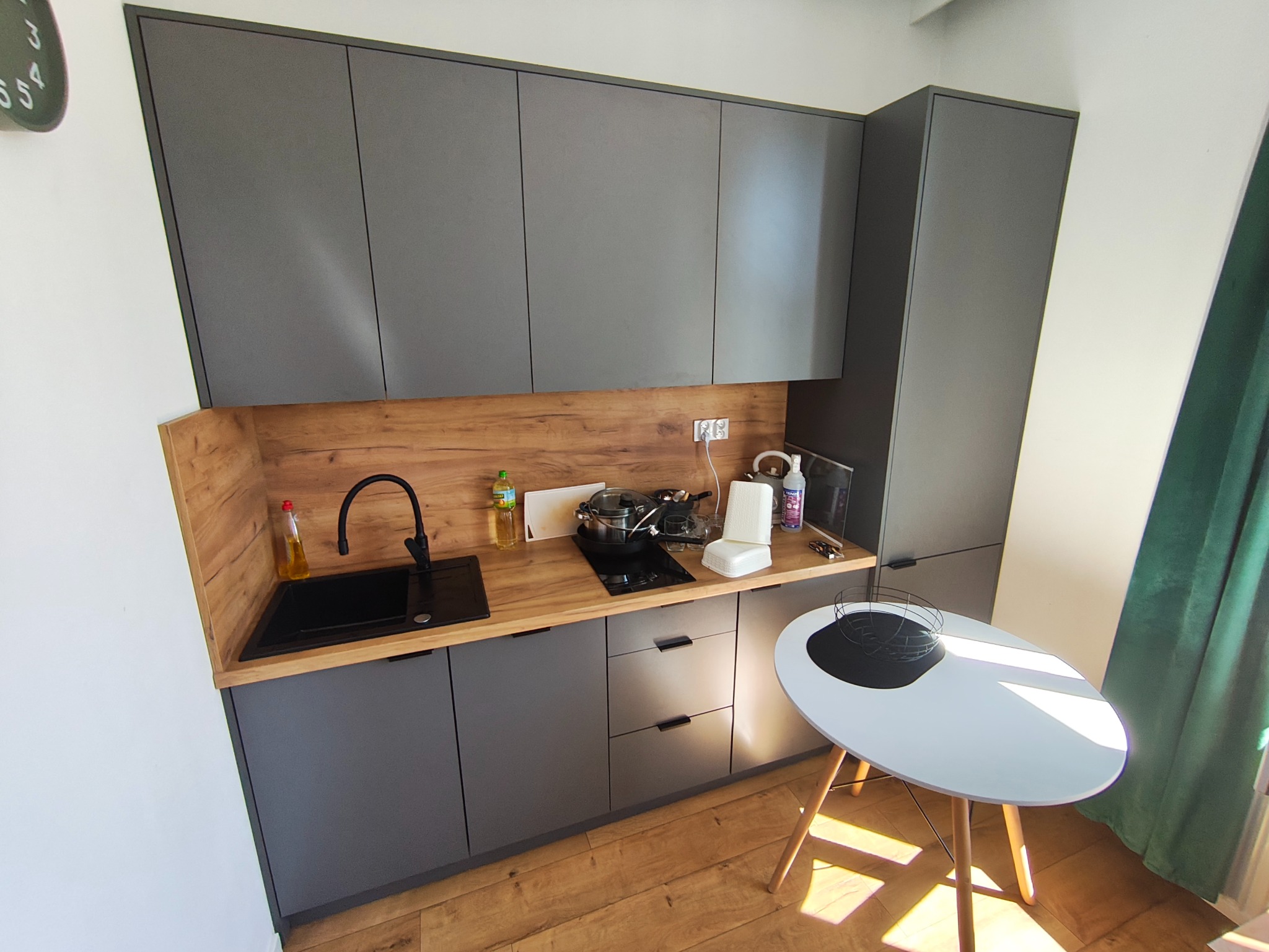 Szare szafki kuchenne z drewnianym blatem, czarny zlew i płyta grzewcza, biały stół z czarną paterą, zielona zasłona w tle. Widok z góry na małą kuchnię.