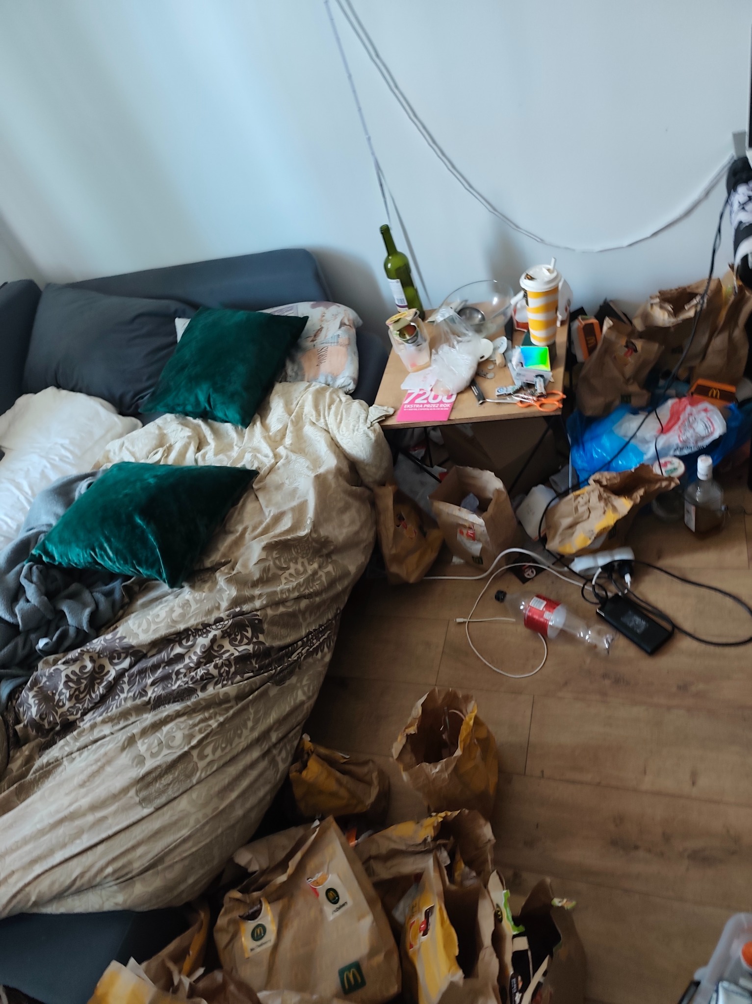 Zaniedbane wnętrze pokoju: rozrzucone śmieci, puste torby po jedzeniu, butelka po winie i niepościelone łóżko z zielonymi poduszkami.