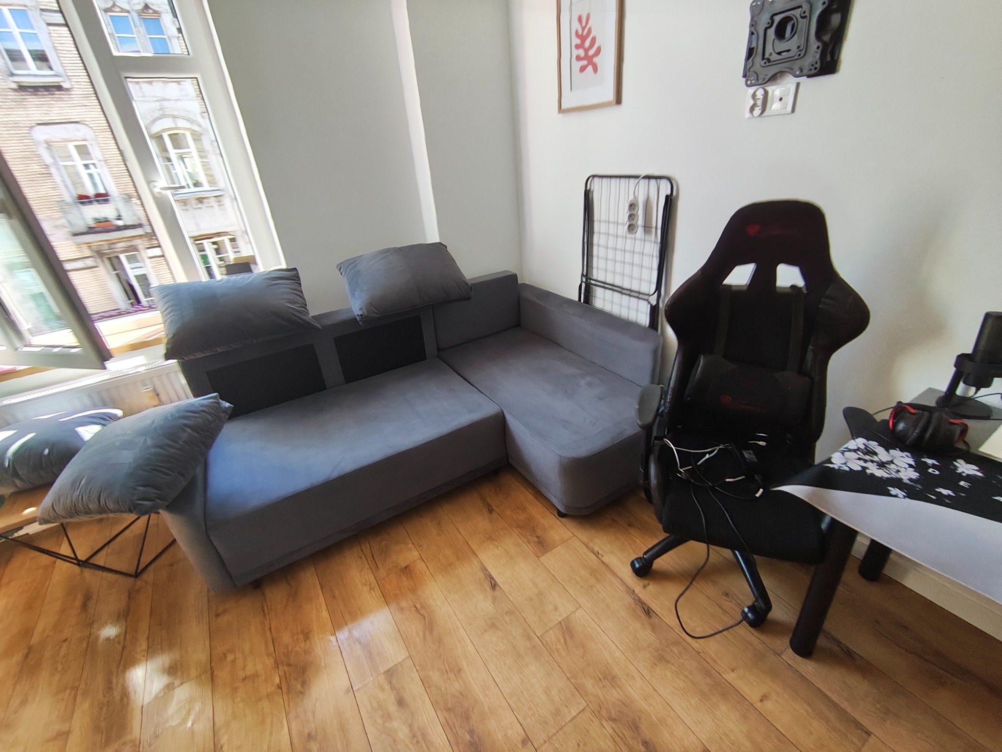 Szary narożnik z poduszkami w jasnym pokoju z drewnianą podłogą w Katowicach. Widok z okna na budynek. Suszarka na pranie i fotel gamingowy w tle.