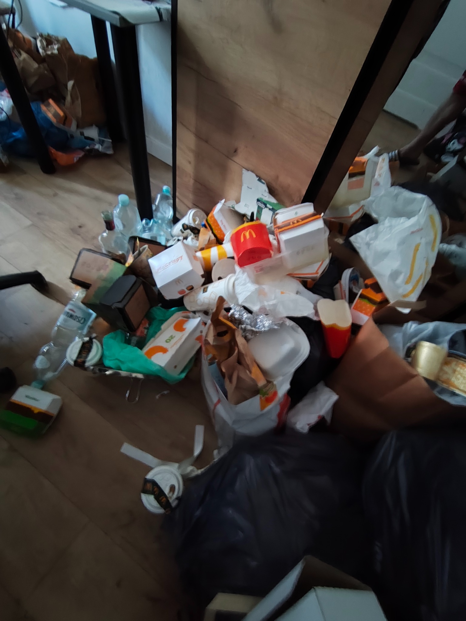 Stos śmieci, w tym pudełka i butelki po napojach, leżący na podłodze w rogu pokoju, blisko nogi stołu. Widoczny czarny worek na śmieci.